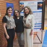 Pesquisadoras diante do pôster sobre a ação seletiva do peptídeo Acro-18 em células de melanoma.